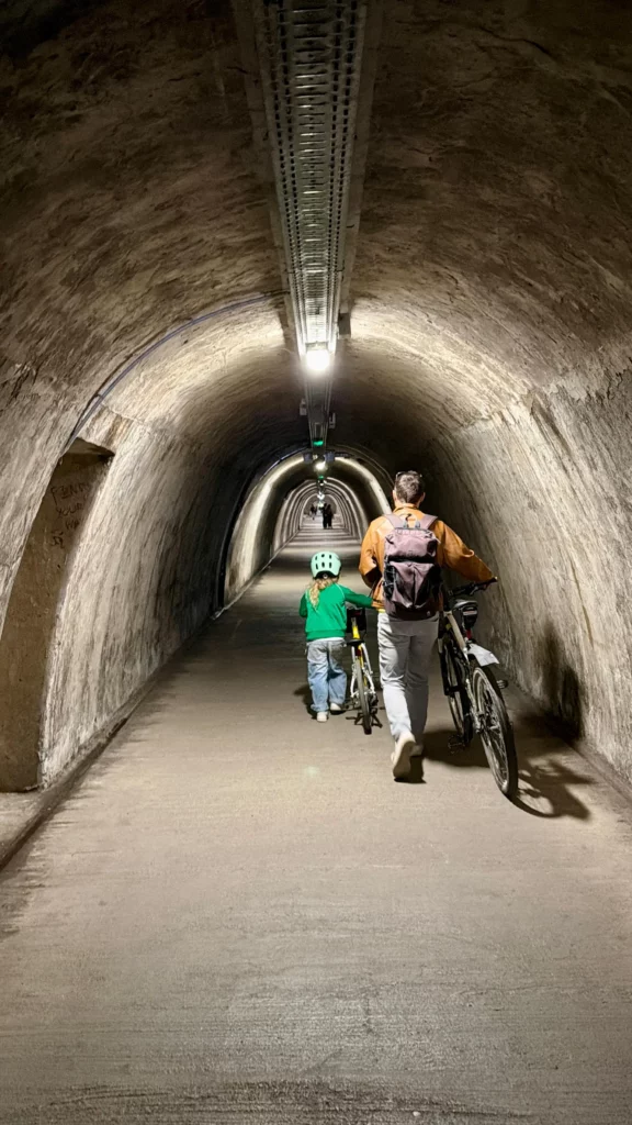 Otac i dijete s dječjom kacigom guraju bicikle hodajući kroz dugi, osvijetljeni pješački tunel Grič u Zagrebu.