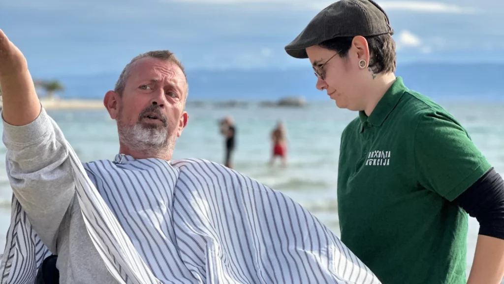 Muškarac s prugastim brijačkim ogrtačem sjedi na plaži i pokazuje rukom u daljinu dok ga osoba u zelenoj majici s logotipom "Bradata aukcija" pažljivo promatra.