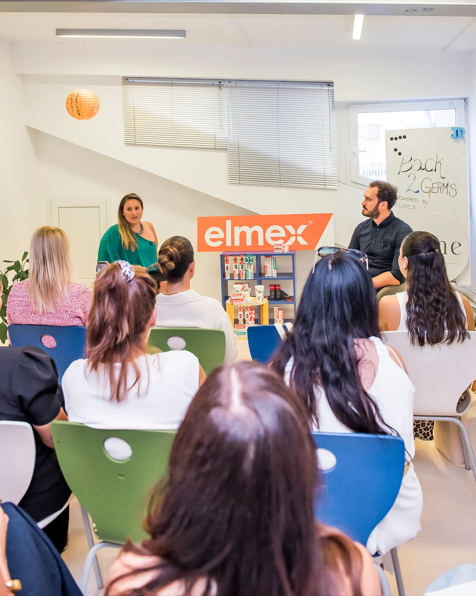 Široki kadar panel rasprave gdje voditeljica i predavač komuniciraju sa sudionicima edukativnog "Back 2 Germs" događaja, smješteni ispred polica s elmex proizvodima.